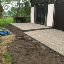 Weles Patryk Macięga - Wyłożony żwirem taras z obrzeżami przy nowoczesnym domu. Niebieski wąż ogrodowy leży obok przygotowanego terenu pod dalsze prace.