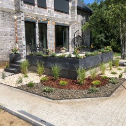 Weles Patryk Macięga - Nowoczesny ogród z podwyższonymi rabatami z czarnych bloczków, ozdobiony trawami, korą i kamieniami. W tle surowy budynek i mężczyzna z taczką.