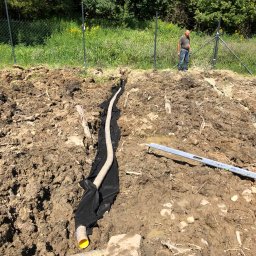 Weles Patryk Macięga - Instalacja drenażu w wykopie, rura drenażowa w geowłókninie, poziomica, pracownik w tle. Teren przygotowany pod przyszły ogród, widoczne ogrodzenie.