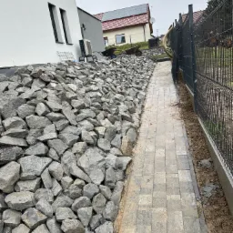 Skarpa z kamienia i ścieżka z kostki brukowej w Jaroszowicach. Ułożony kamień na skarpie, obok ścieżka, w tle dom i ogrodzenie panelowe. Widok na posesję.