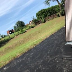 Przygotowanie terenu pod trawnik z rolki: rozłożona czarna agrowłóknina na ziemi, w tle zielona trawa i zabudowa jednorodzinna, błękitne niebo.