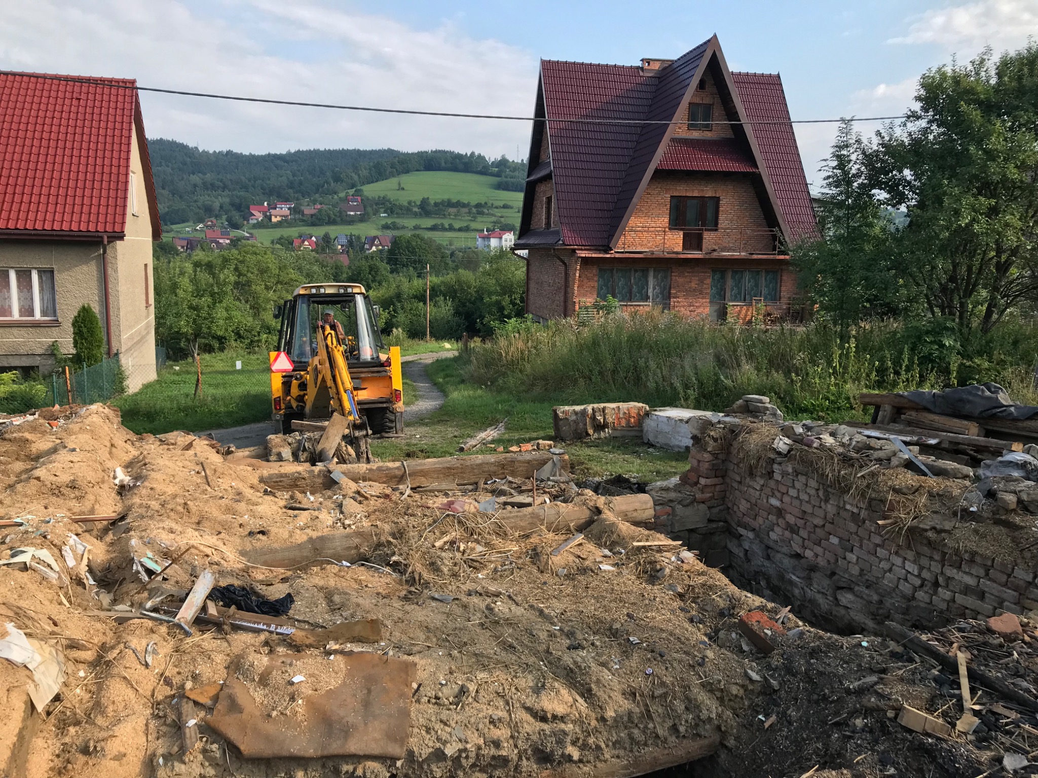 Żółta koparka na działce po rozbiórce, w tle domy i wzgórza. Gruz i ziemia na pierwszym planie. Prace ziemne w toku, widok na posesję w słoneczny dzień.