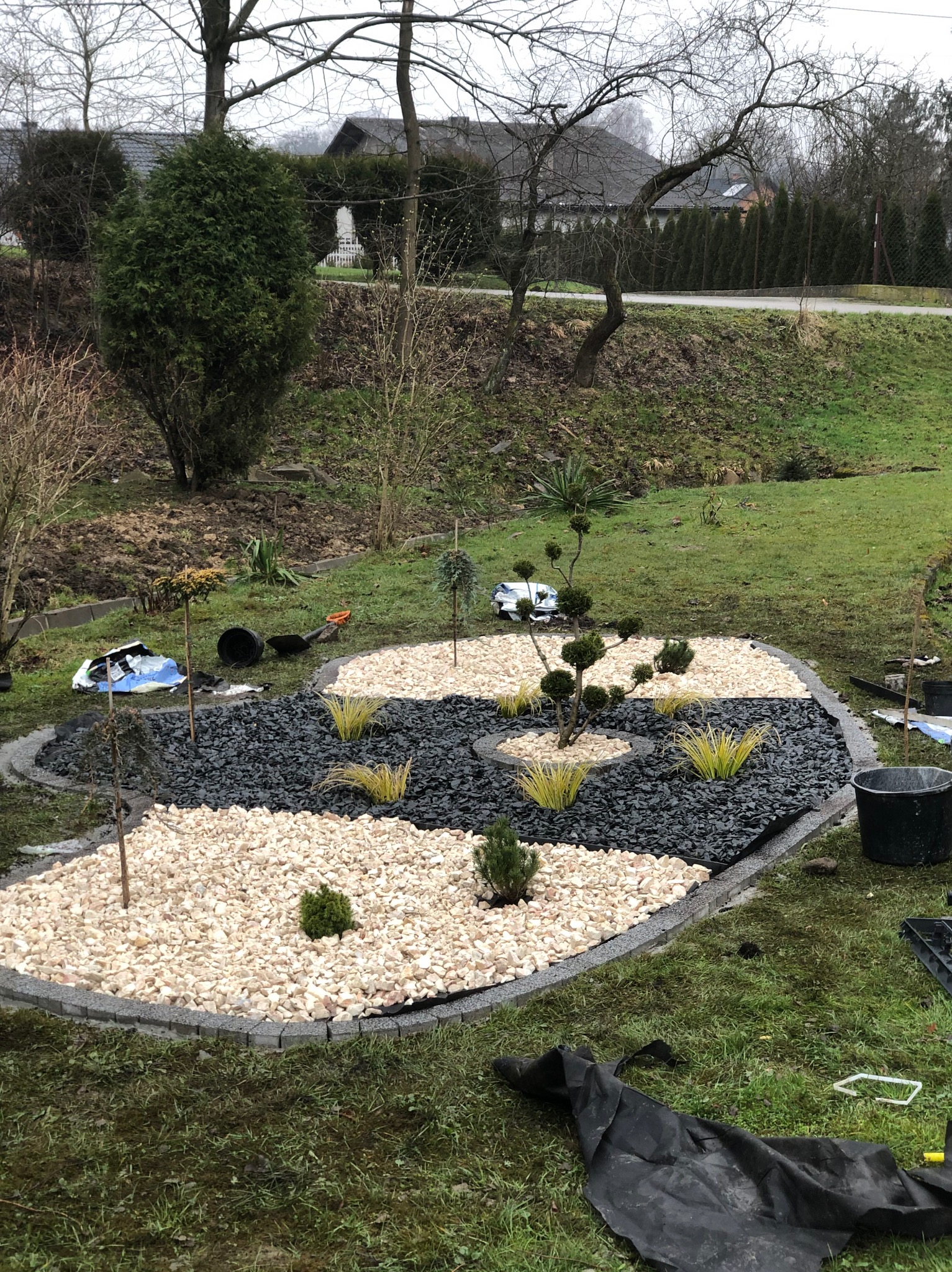 Aranżacja ogrodu z białym i czarnym grysem, obrzeża z kostki, niskie krzewy ozdobne. Na trawie widoczne narzędzia ogrodnicze i resztki materiałów.