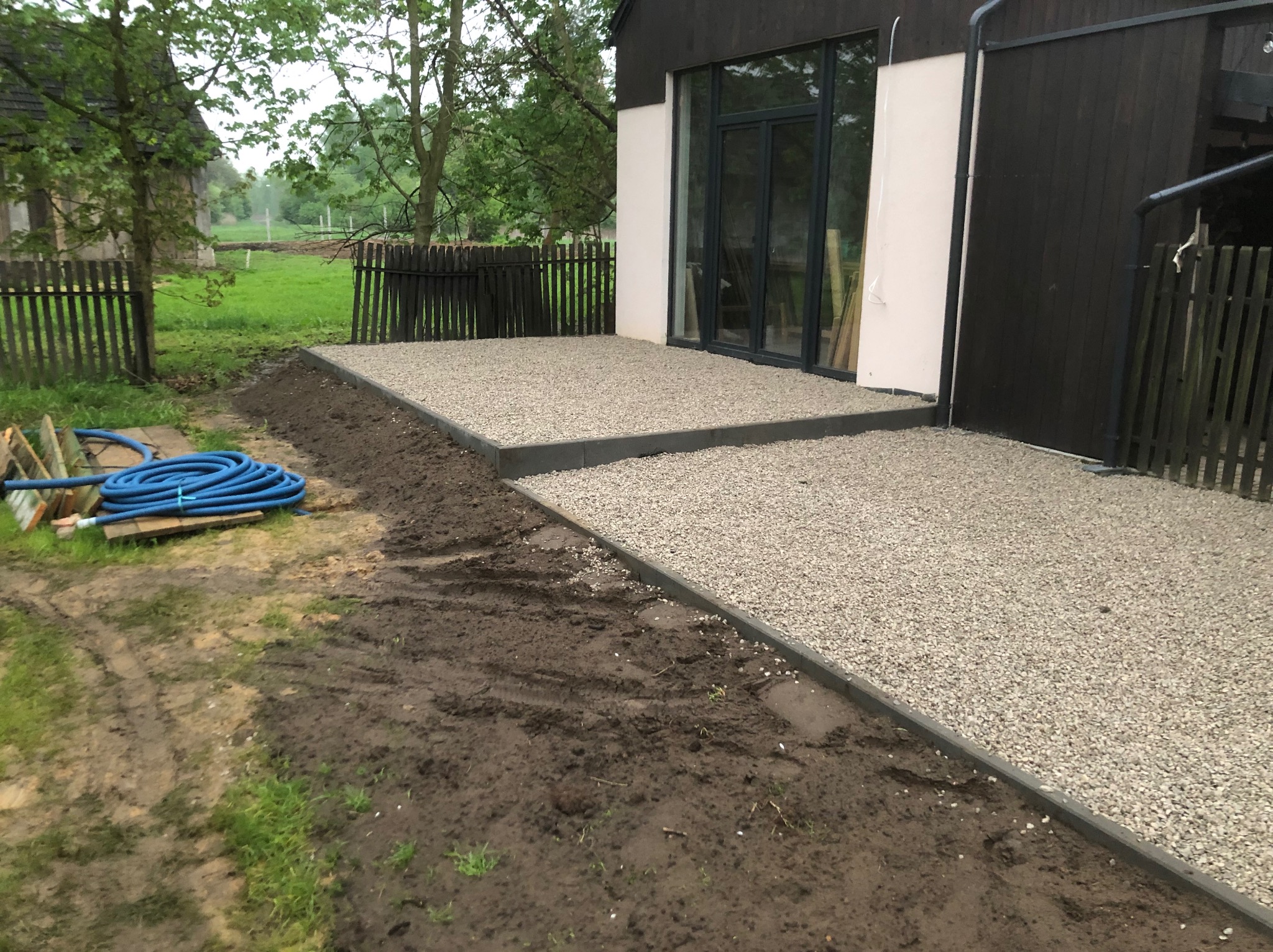 Wyłożony żwirem taras z obrzeżami przy nowoczesnym domu. Niebieski wąż ogrodowy leży obok przygotowanego terenu pod dalsze prace.