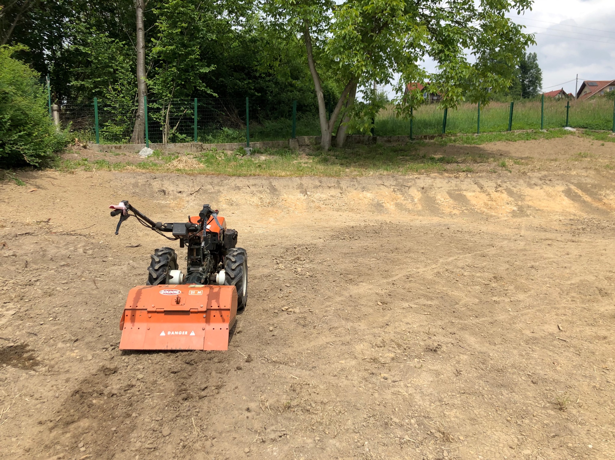 Pomarańczowy glebogryz na przygotowanym podłożu, widoczne drzewa i trawa w tle. Teren wyrównany, gotowy do dalszych prac ogrodniczych.