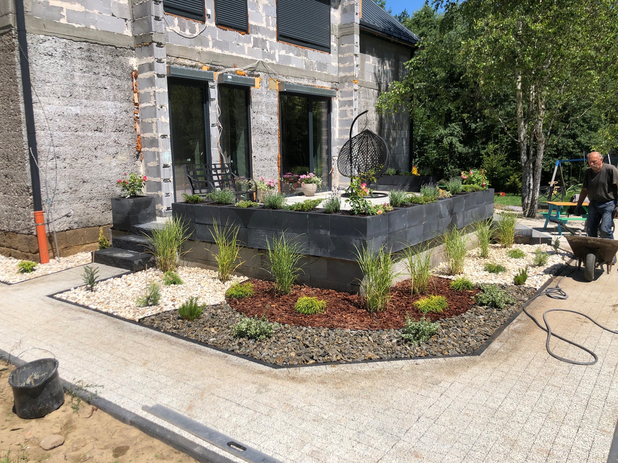 Nowoczesny ogród z podwyższonymi rabatami z czarnych bloczków, ozdobiony trawami, korą i kamieniami. W tle surowy budynek i mężczyzna z taczką.