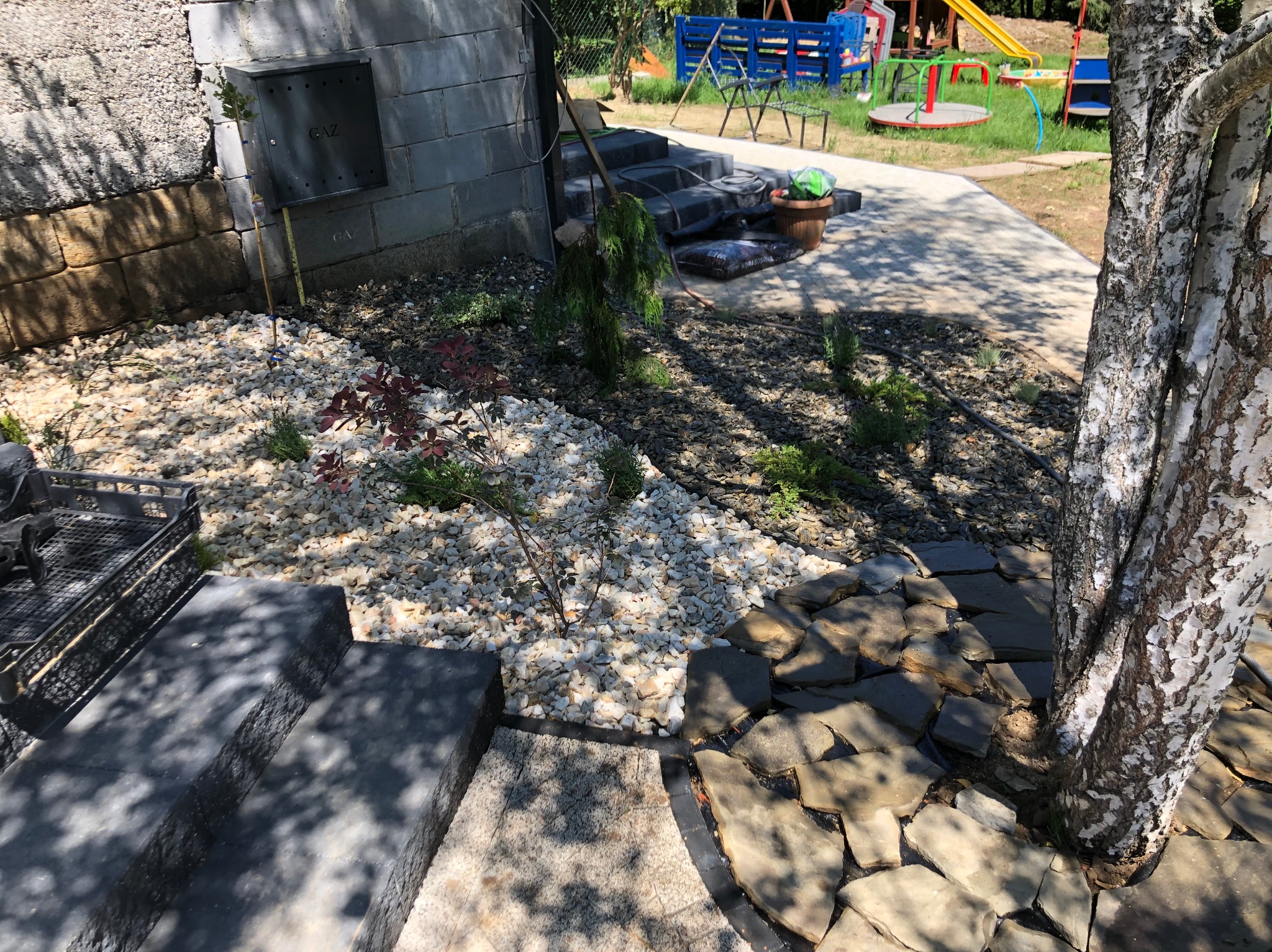Ogród z białym żwirem, kamiennymi płytami wokół brzozy i schodami. W tle plac zabaw i budynek z instalacją gazową. Estetyczne zagospodarowanie przestrzeni.