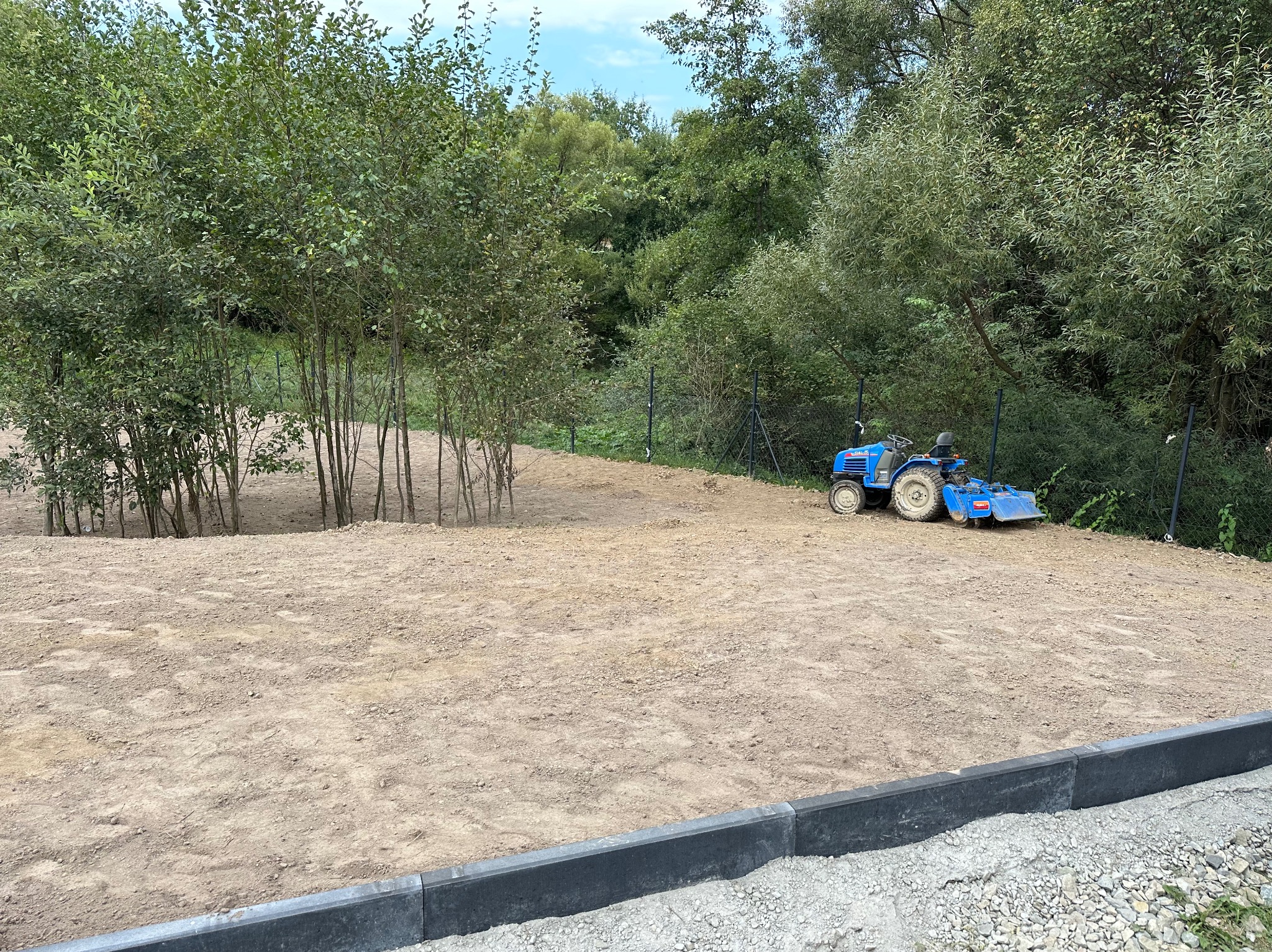 Przygotowany teren pod trawnik z obrzeżami, mały traktorek ogrodowy w tle, młode drzewka w lewym rogu. Niwelacja, wyrównanie ziemi.