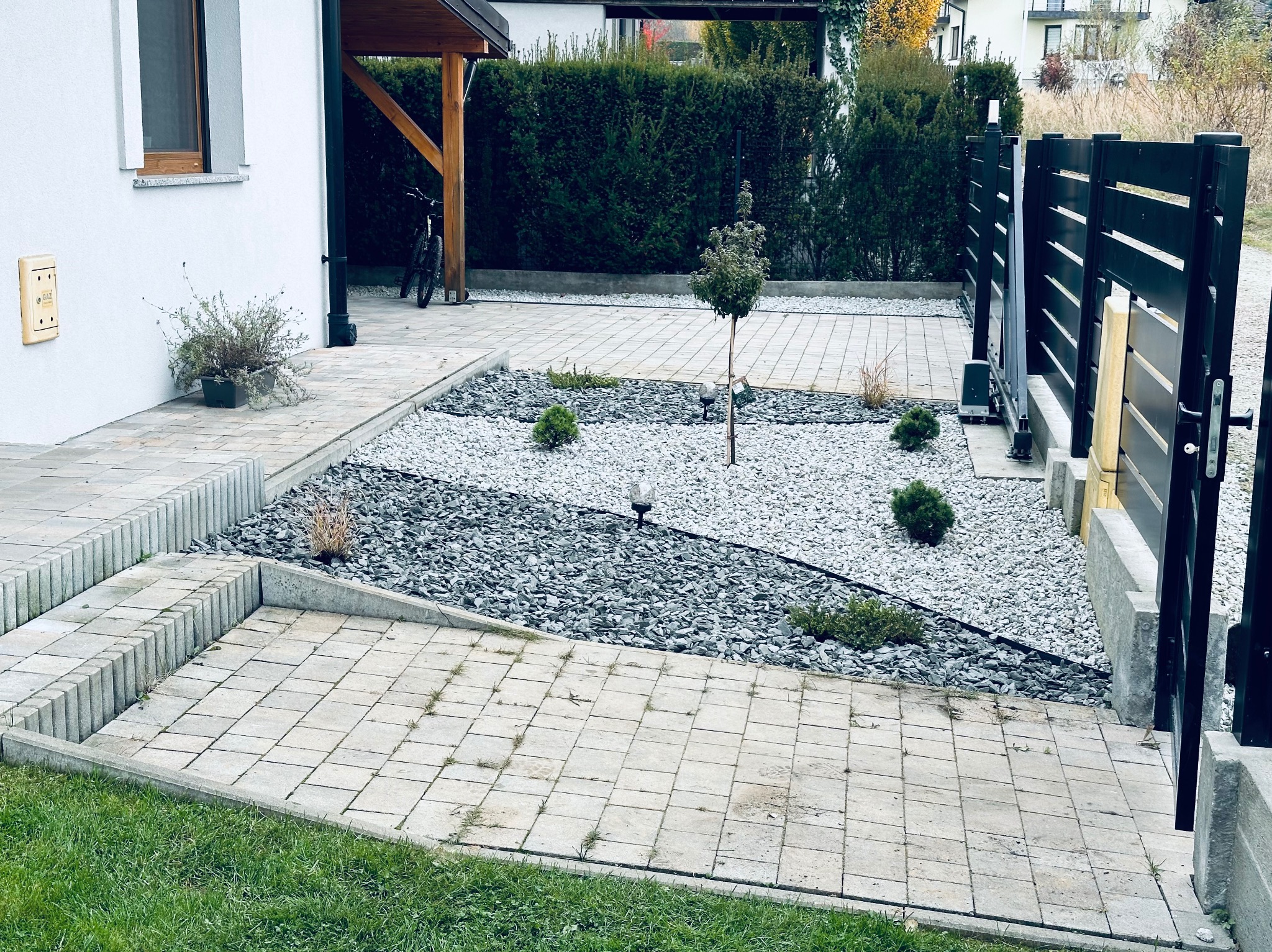 Nowoczesny ogród z jasną kostką brukową, szarym żwirem i niskimi roślinami ozdobnymi. W tle żywopłot i fragment domu z drewnianą wiatą.