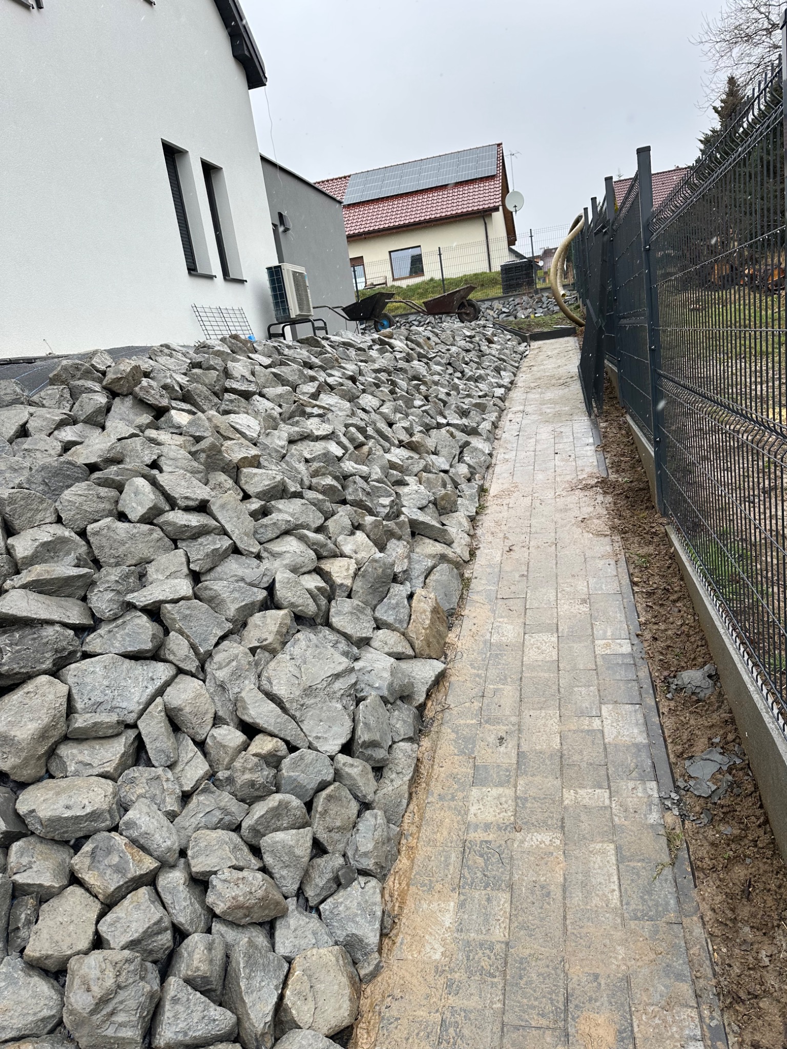 Skarpa z kamienia i ścieżka z kostki brukowej w Jaroszowicach. Ułożony kamień na skarpie, obok ścieżka, w tle dom i ogrodzenie panelowe. Widok na posesję.