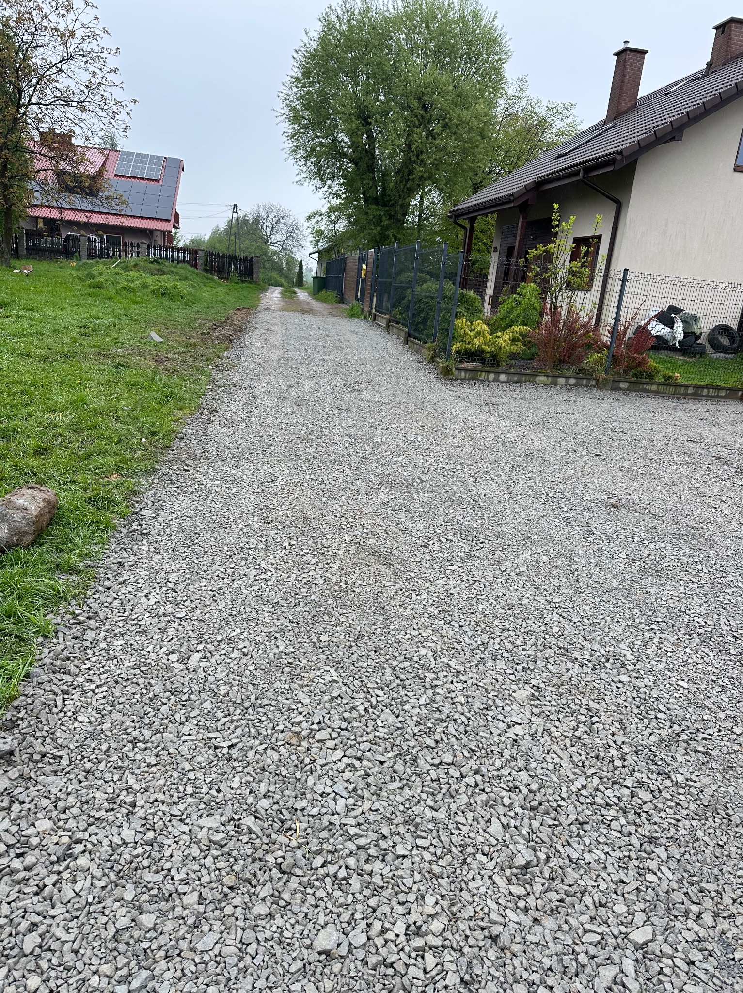 Szutrowa droga wjazdowa do posesji z domami w tle, widok z perspektywy pieszego. Świeżo wysypany szuter, trawa po bokach, pochmurne niebo.