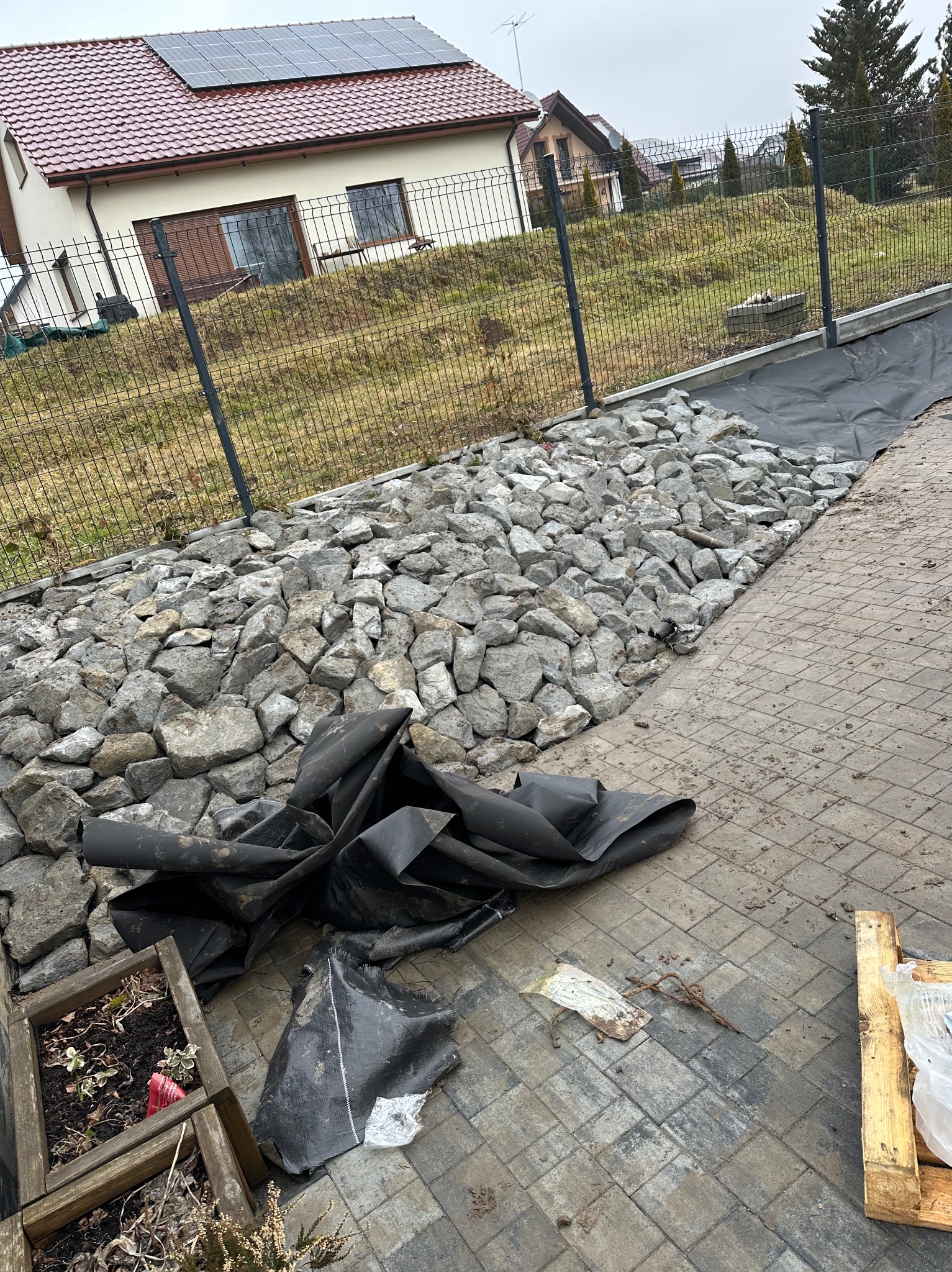 Układanie kamieni i czarnej agrowłókniny w ogrodzie przy domu jednorodzinnym, widoczne narzędzia i materiały, etap prac ziemnych.