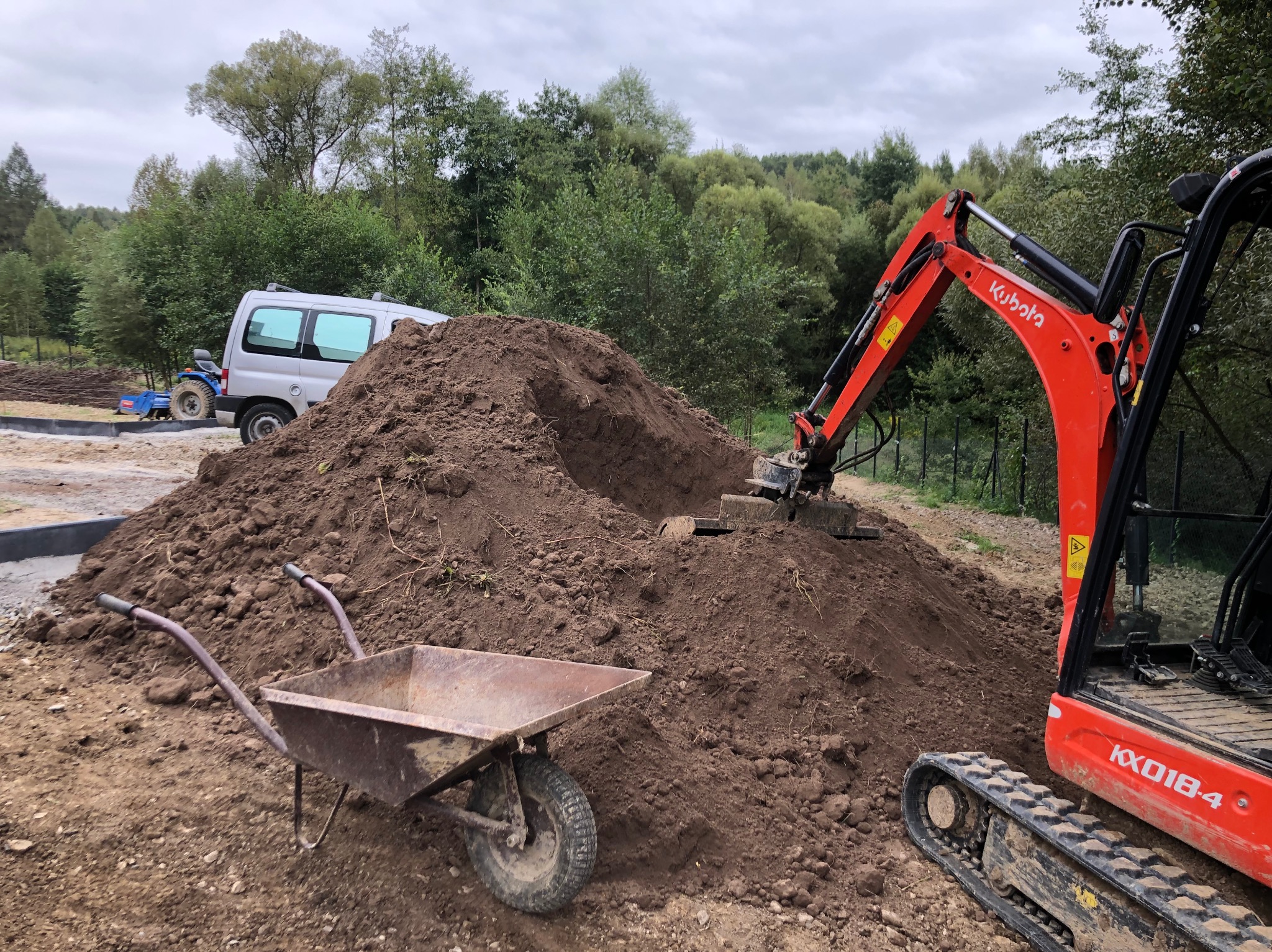 Koparka Kubota przy pryzmie ziemi, taczka obok. W tle auto i mały traktor, teren zielony. Niwelacja terenu w toku, przygotowanie pod ogród. Prace ziemne.