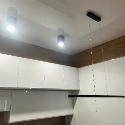 Nowoczesna kuchnia z białymi szafkami i oświetleniem: dwa cylindryczne reflektory i długa, pozioma lampa wisząca. Drewnopodobne wykończenie nad szafkami.