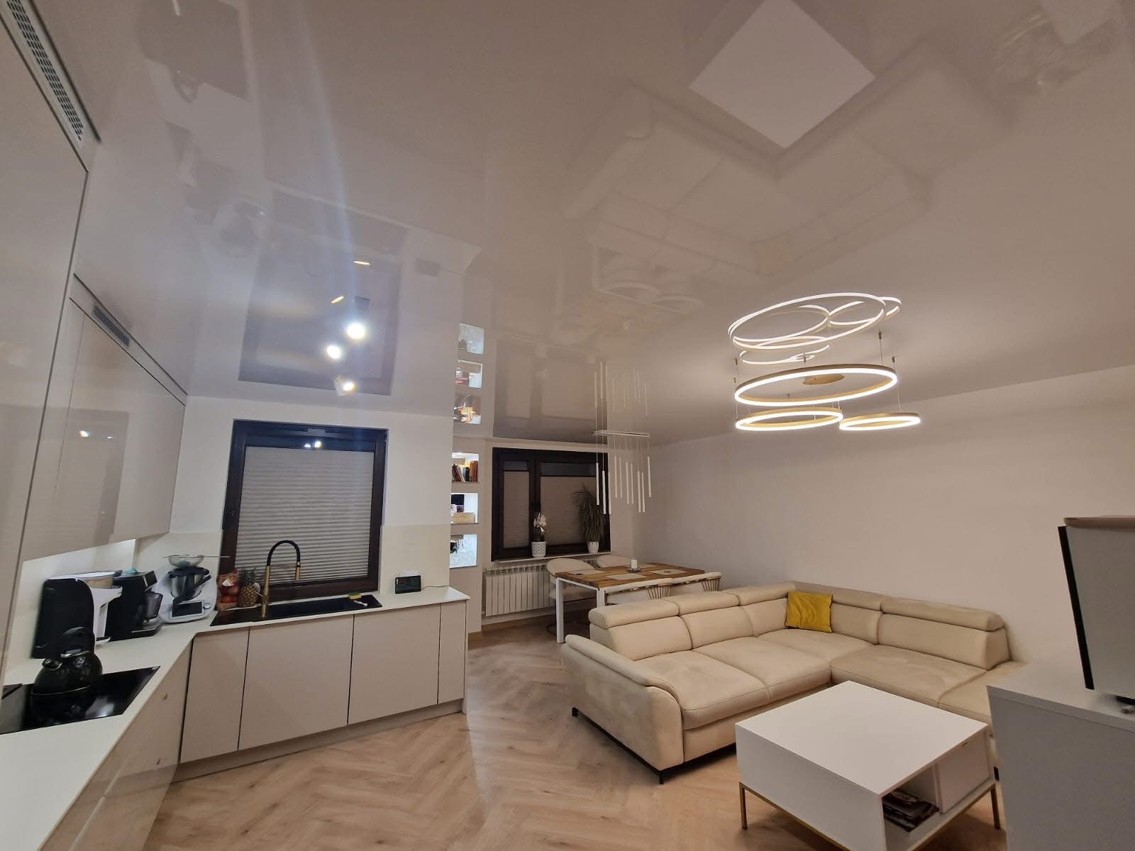 Błyszczący sufit napinany z wbudowanym oświetleniem w nowoczesnym salonie z aneksem kuchennym, widok z dołu, podkreślający gładką powierzchnię i odbijające światło właściwości.