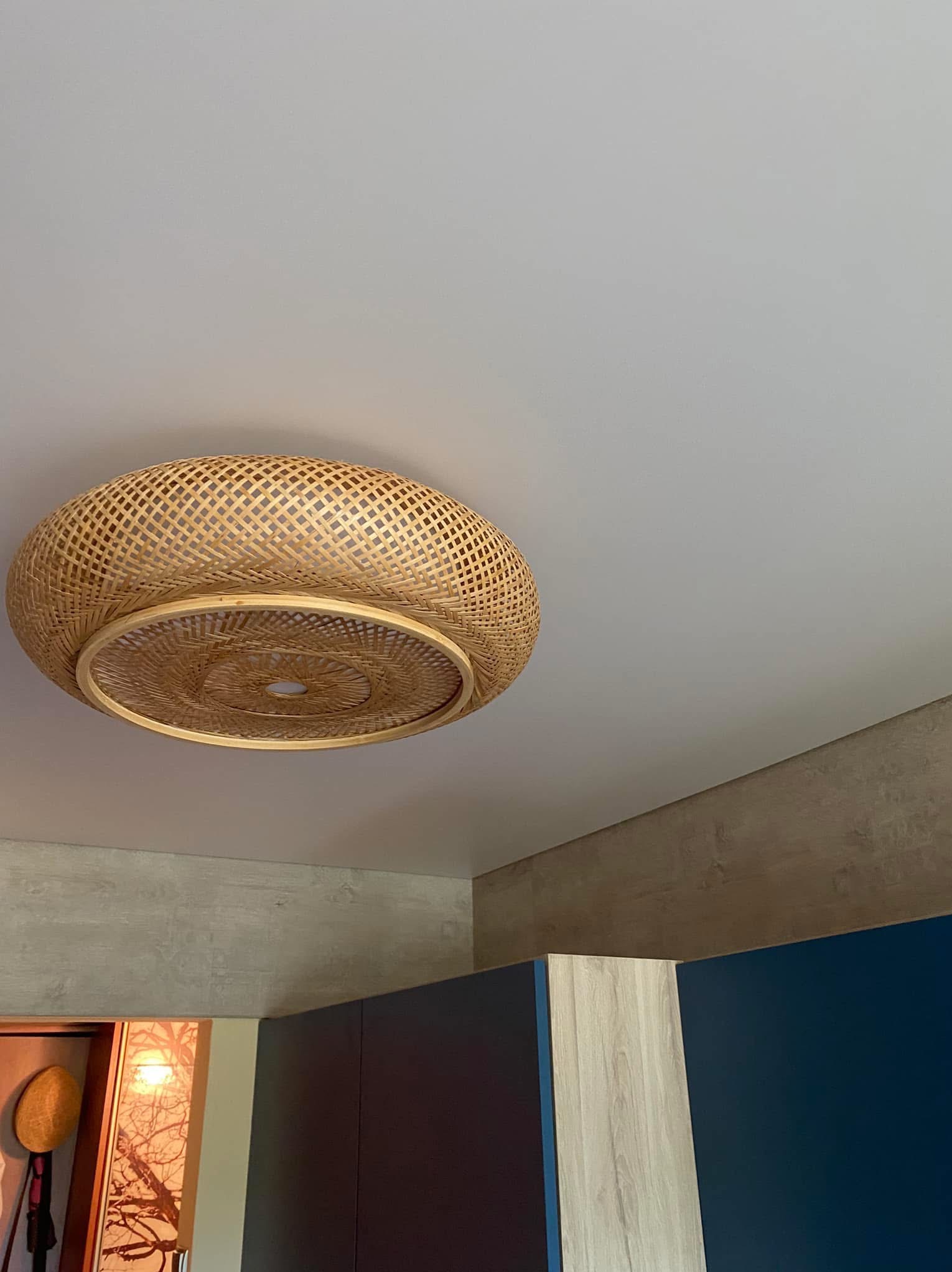 Okrągła, pleciona lampa sufitowa z bambusa, zamontowana na gładkim, białym suficie. Widoczne fragmenty ściany i zabudowy meblowej w tle.