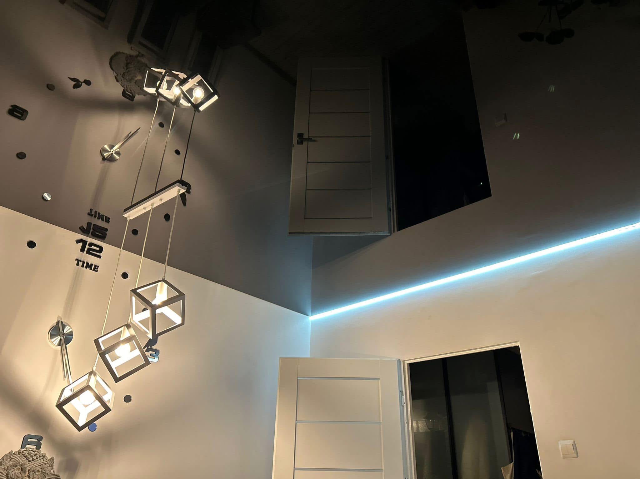 Nowoczesny pokój z ciemnym, błyszczącym sufitem, oświetlonym listwą LED i dekoracyjnym zegarem ściennym oraz lampą w kształcie kostek.