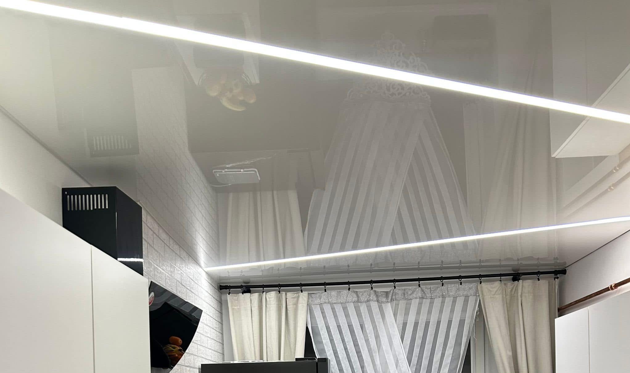 Błyszczący, biały sufit napinany z oświetleniem LED, odbijający fragment kuchni z okapem i zasłonami. Widoczna faktura ściany z cegieł.