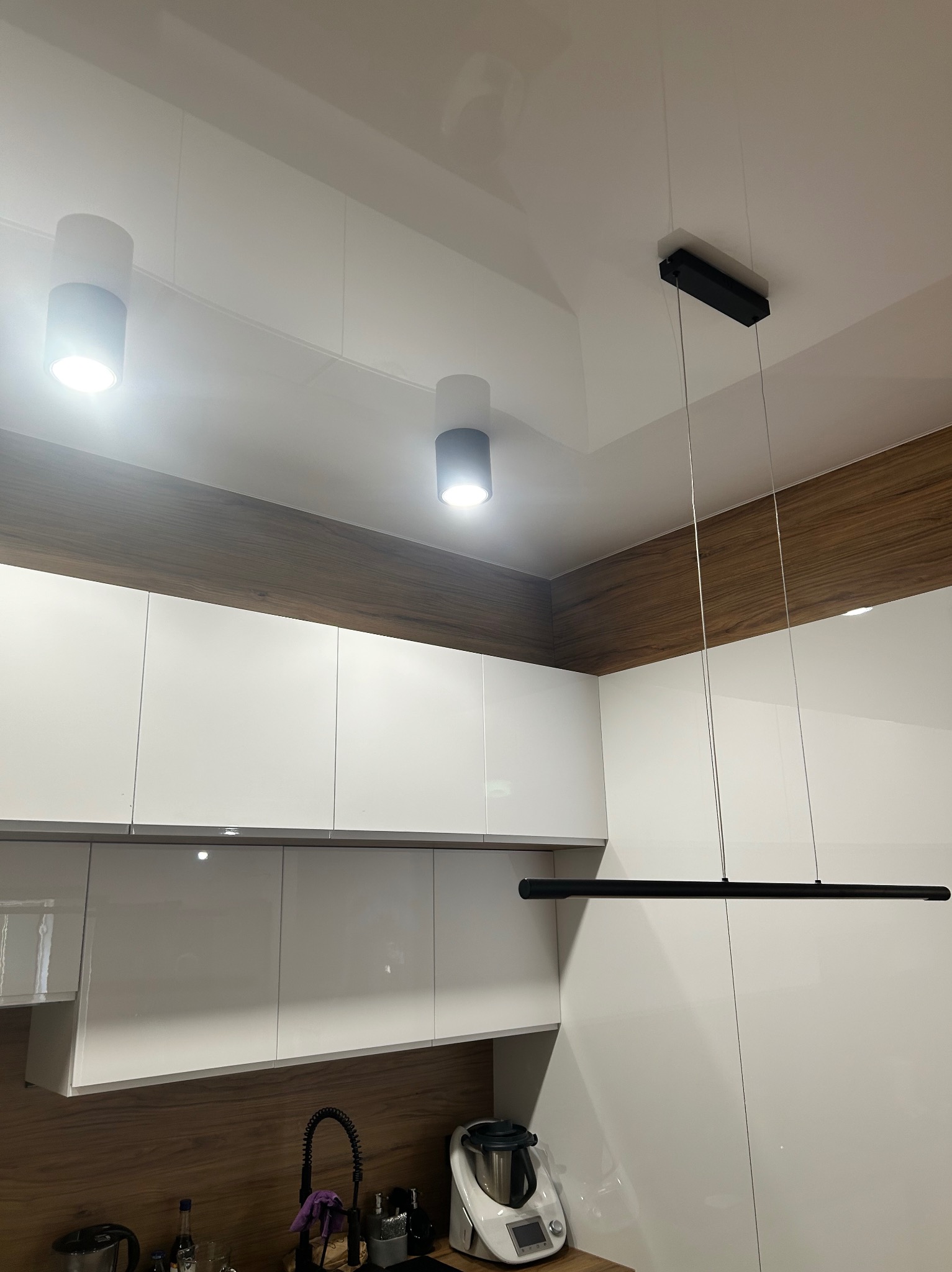 Nowoczesna kuchnia z białymi szafkami i oświetleniem: dwa cylindryczne reflektory i długa, pozioma lampa wisząca. Drewnopodobne wykończenie nad szafkami.