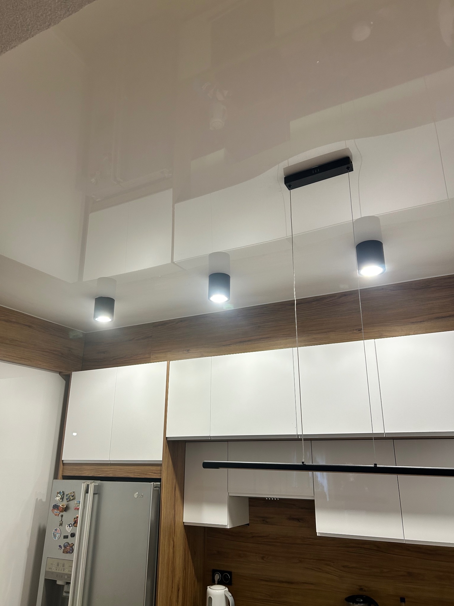 Biały, błyszczący sufit z trzema okrągłymi lampami i jedną podłużną, zwisającą nad zabudową kuchenną z białymi szafkami i drewnopodobnymi panelami. Nowoczesne oświetlenie.