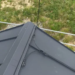 Czarna instalacja odgromowa zamontowana na szarym metalowym dachu, widok z góry na tle zieleni i rusztowania.