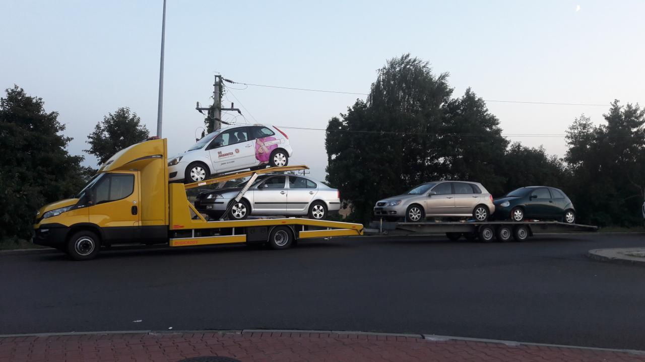 Żółta ciężarówka z platformą transportową przewozi trzy samochody osobowe na dwóch poziomach oraz jeden samochód na przyczepie, w tle drzewa i słup energetyczny.