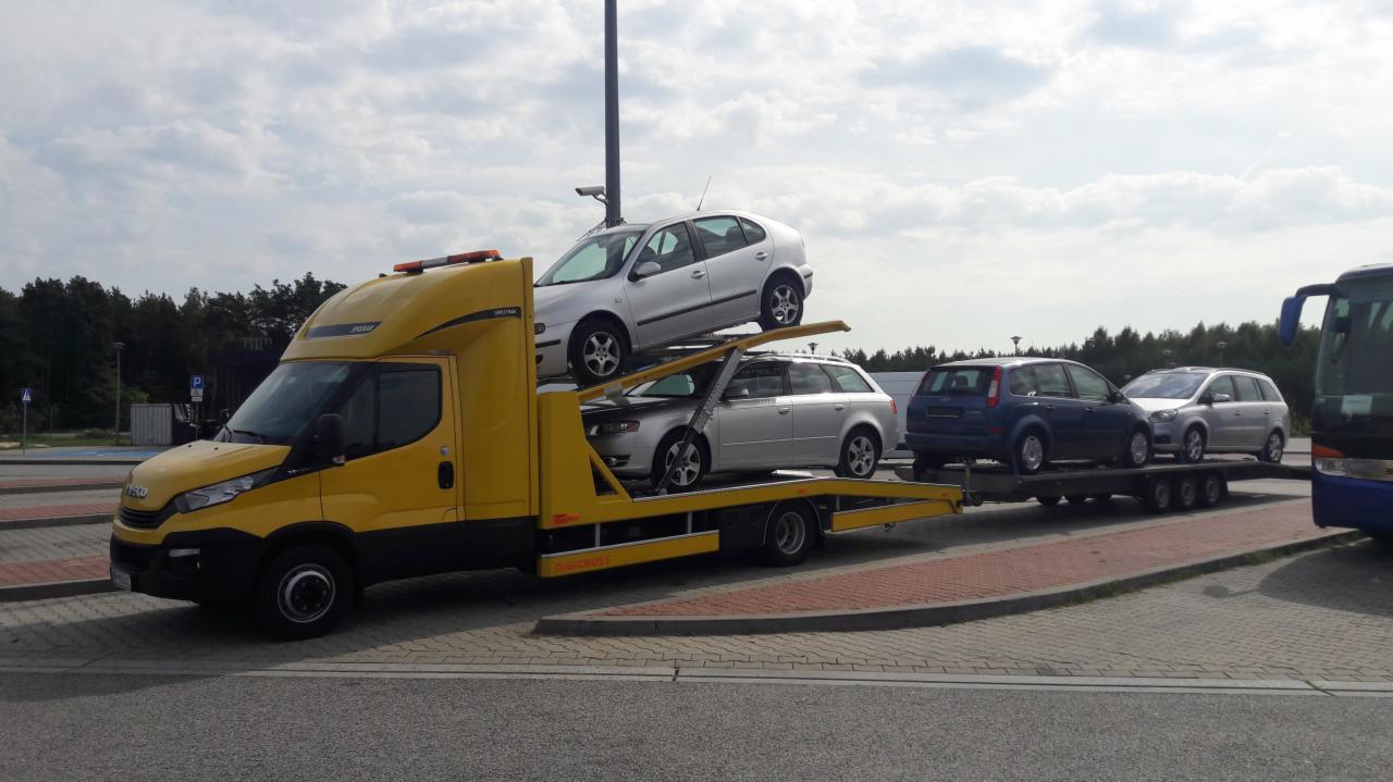 Żółta ciężarówka z platformą transportową przewozi trzy samochody osobowe, stojąca na parkingu z widocznym autobusem w tle.