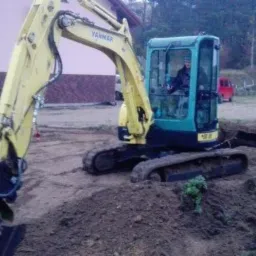 Żółto-turkusowa mini koparka Yanmar z operatorem w kabinie, pracująca na nierównym terenie z pryzmami ziemi, w tle budynek i zalesiony teren.