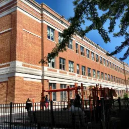 Wielopiętrowy budynek z czerwonej cegły z białymi elementami dekoracyjnymi, widoczny plac zabaw i fragment drzewa z prawej strony.