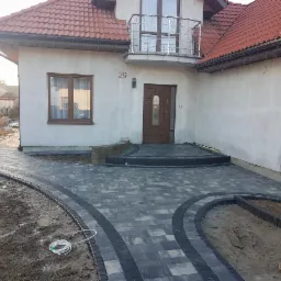 Nowo ułożona, szara kostka brukowa z ciemniejszym obramowaniem, prowadząca do domu z brązowymi drzwiami wejściowymi i numerem 29, widoczna w nieukończonym ogrodzie.