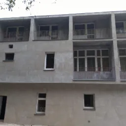 Szary budynek wielorodzinny w trakcie prac wykończeniowych elewacji, widoczne balkony z metalowymi balustradami i okna o różnych rozmiarach, otwarte drzwi wejściowe na parterze.