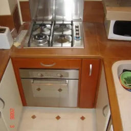 Kuchenka gazowa z czterema palnikami i piekarnikiem w zabudowie z brązowymi frontami, białe płytki podłogowe z brązowymi akcentami, widoczne akcesoria kuchenne i pudełko z elektroniką.