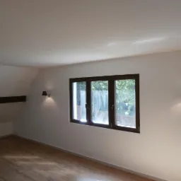 Poddasze po remoncie z drewnianą podłogą, trzema oknami w ciemnych ramach i dwoma czarnymi kinkietami po bokach okna.