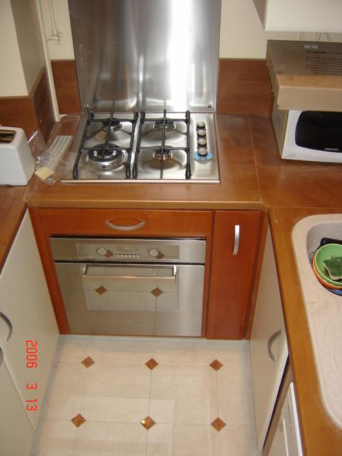 Kuchenka gazowa z czterema palnikami i piekarnikiem w zabudowie z brązowymi frontami, białe płytki podłogowe z brązowymi akcentami, widoczne akcesoria kuchenne i pudełko z elektroniką.