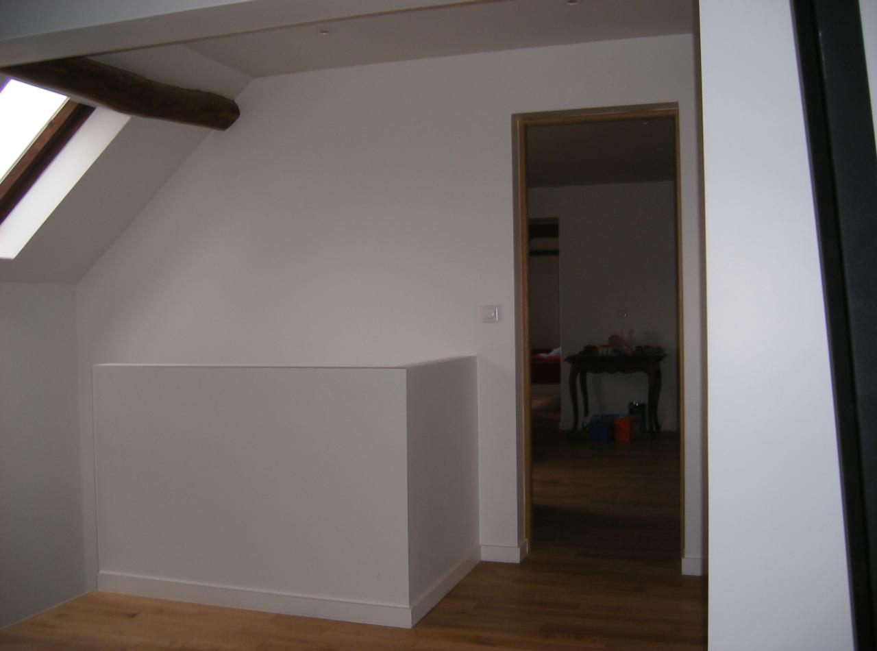 Poddasze po remoncie z drewnianą belką stropową, białą ścianą działową i widokiem na pokój przez drewnianą futrynę drzwiową.