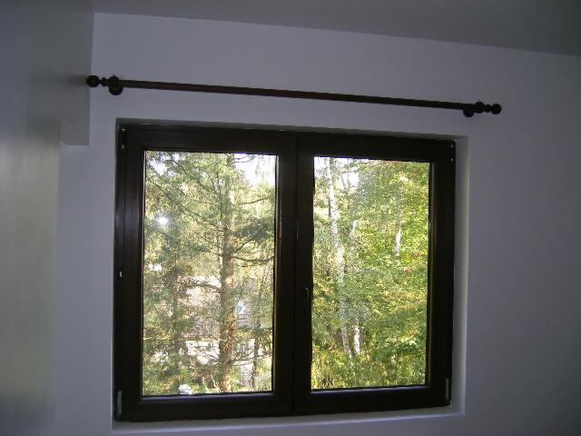 Dwuskrzydłowe okno z brązową ramą, widok na drzewa w tle, zamontowany karnisz z ozdobnymi zakończeniami.