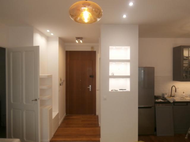 Wnętrze mieszkania po remoncie: przedpokój z drewnianą podłogą przechodzący w kuchnię, widoczna zabudowana wnęka z półkami, oświetlenie sufitowe i wisząca lampa. Kuchnia z szarą zabudową i srebrną...