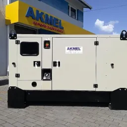 Agregat prądotwórczy marki Akmel w obudowie, stojący na kostce brukowej, z widocznym logo producenta na obudowie i szyldem firmy w tle.