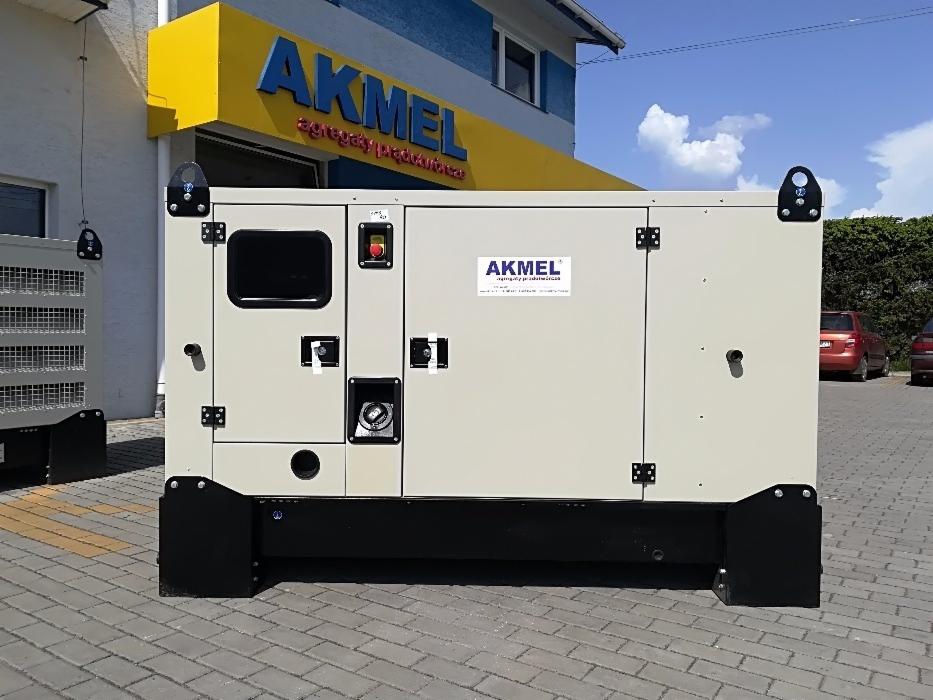 Agregat prądotwórczy marki Akmel w obudowie, stojący na kostce brukowej, z widocznym logo producenta na obudowie i szyldem firmy w tle.