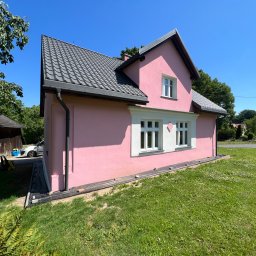 Docieplenia&elewacje - Różowy dom jednorodzinny z szarą dachówką i białymi okiennicami, widok z boku, świeżo pomalowana elewacja w słoneczny dzień.