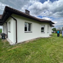 Docieplenia&elewacje - Biały dom jednorodzinny z brązową dachówką i rynnami, widoczny pracownik przy elewacji i drugi z panelem solarnym na trawniku, pochmurne niebo.