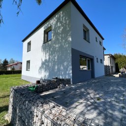 Docieplenia&elewacje - Ocieplony dom jednorodzinny z nową elewacją w kolorze białym i szarym, widoczny z boku, z kamienną gabionową ścianą i wybrukowanym podjazdem, w słoneczny dzień.