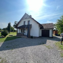 Docieplenia&elewacje - Jasny, dwukondygnacyjny dom z garażem, białymi ścianami i brązowym dachem, widziany w słoneczny dzień z podjazdu wysypanego żwirem; w tle zieleń drzew, a obok domu stoi mężczyzna ze skuterem.