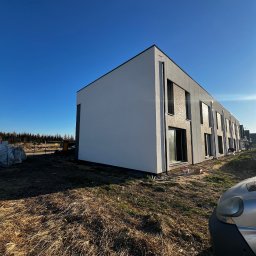 Docieplenia&elewacje - Nowoczesny budynek szeregowy z białą elewacją i dekoracyjnymi panelami, widoczny w surowym krajobrazie z elementami budowlanymi w tle, sfotografowany w słoneczny dzień.