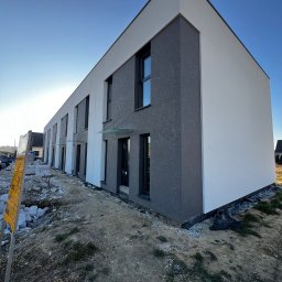 Docieplenia&elewacje - Nowoczesny budynek wielorodzinny z elewacją w kolorze białym i szarym, widoczne szklane zadaszenia nad wejściami, teren wokół budynku w trakcie prac wykończeniowych.