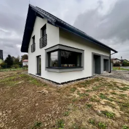 Nowoczesny, biały dom z szarą stolarką i dachem w Skoczowie, z widocznym oknem narożnym i balkonami, na tle pochmurnego nieba i przygotowanego terenu pod ogród.