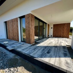 Docieplenia&elewacje - Nowoczesny taras z szarych płyt betonowych i drewnianą elewacją pionową, widoczne przygotowania do dalszych prac wykończeniowych, kubełek z zaprawą.