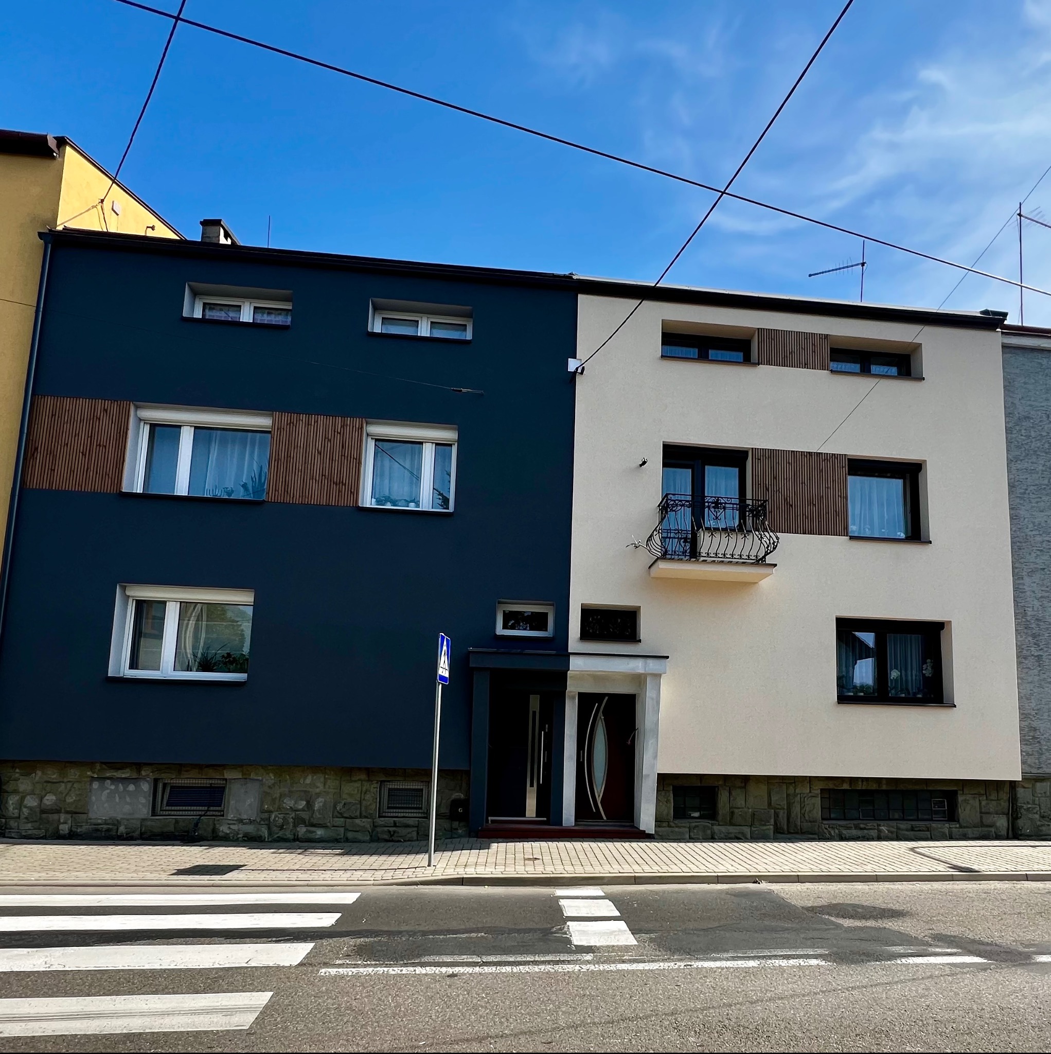 Dwukolorowa fasada budynku z ciemnoniebieską i kremową częścią, ozdobiona drewnianymi panelami, balkonem z kutą balustradą i widocznym przejściem dla pieszych.