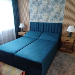 Bauel Tomasz Iskrzak - Nowoczesna sypialnia z turkusowym łóżkiem kontynentalnym, drewnianymi lamelami na ścianie i wzorzystą tapetą. Widoczne szafki nocne z lampkami.