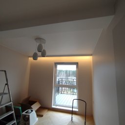 Bauel Tomasz Iskrzak - Jasne wnętrze po remoncie z oświetleniem LED podsufitowym, drabina i wózek transportowy. Widoczny parkiet w jodełkę i okno z widokiem na balkon.